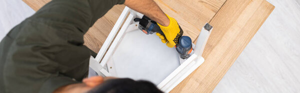 Overhead view of young man repairing chair with electric screwdriver at home, banner  