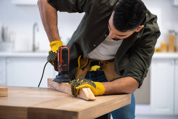 Muslim man with tool belt sanding wooden board at home 