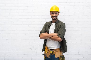Cheerful arabian repairman in hardhat crossing arms at home 