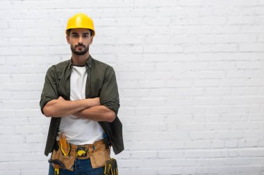 Arabian handyman in helmet and tool belt looking at camera at home 