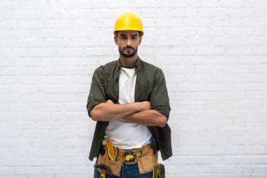 Young muslim handyman in helmet and tool belt looking at camera at home 
