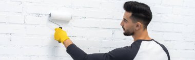 Side view of arabian man coloring white brick wall at home, banner 