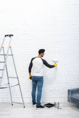 Side view of muslim man in gloves coloring brick wall with roller at home 