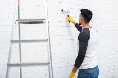 Arabian man in gloves coloring wall near ladder at home 