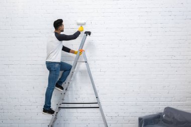 Arabian man in gloves coloring wall while standing on ladder at home 