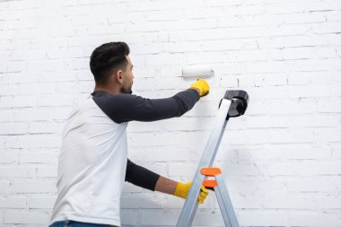 Young muslim man painting wall with roller while standing on ladder in living room 