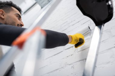 Low angle view of muslim man painting brick wall near blurred ladder at home