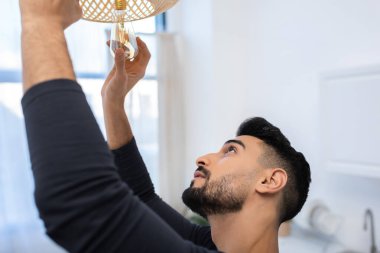 Side view of arabian man changing lightbulb in chandelier in kitchen 