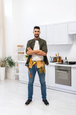 Cheerful muslim man with tool belt crossing arms in kitchen 