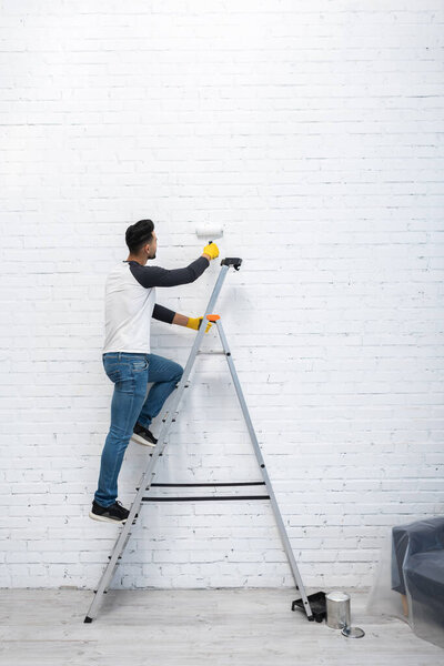 Arabian man coloring brick wall while standing on ladder in living room 