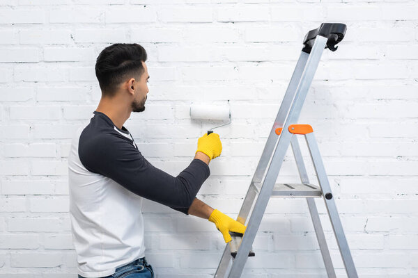 Young arabian man painting white brick wall and holding ladder at home 