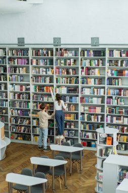 back view of teenage friends choosing books on racks in library