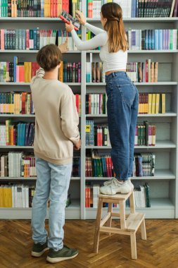 back view of teenage girl standing on step stool near friend and choosing books on shelves