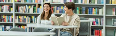 cheerful teenage girl sitting with book on head near laughing friend in reading room, banner