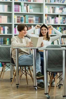 cheerful teen girl covering head with book near smiling friend reading in library