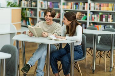 smiling guy reading book near laughing teenage girl in library