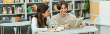 cheerful student reading book near smiling teenage girl in library, banner