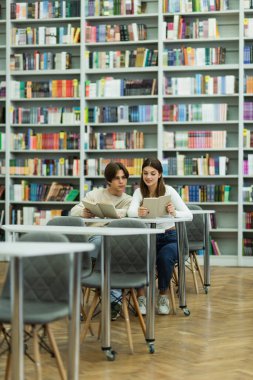 smiling teenage girl reading book near friend and racks in library