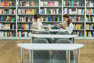 teenage girl with book pointing with hand while talking to friend in library
