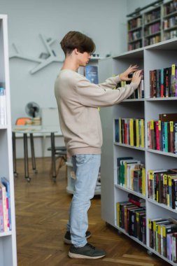 side view of teenage student choosing books on racks in library