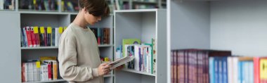 teenager reading book while standing near racks in library, banner