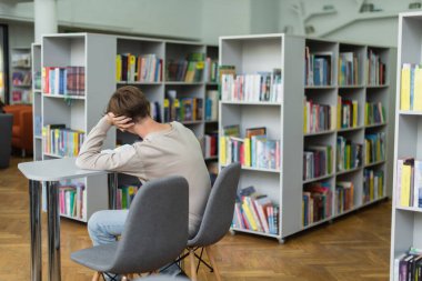 back view of teenage student sitting in library reading room near bookshelves