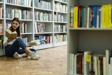 teenage student choosing books while sitting on floor in library