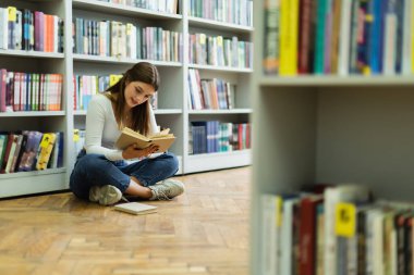 teen girl sitting on floor with crossed legs and reading book in library on blurred foreground