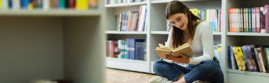 teen student sitting on floor in library and reading book on blurred foreground, banner
