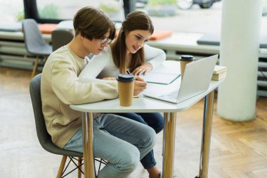 teenage girl smiling near laptop and guy writing in library reading room