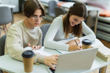 student using laptop near happy teenage girl writing in notebook in library