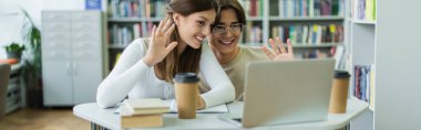 smiling teenage students waving hands during video chat on laptop in library, banner