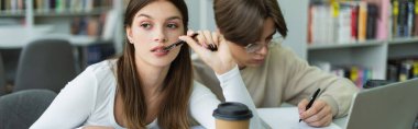 thoughtful teenage girl with pen thinking near writing friend and laptop in library, banner