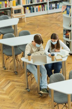 high angle view of teenagers in medical masks writing near laptop and books in library 