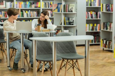 smiling teenage girl talking to friend near laptop in library reading room