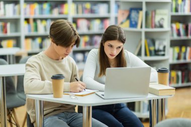 student writing in notebook near teenage girl looking at laptop in library