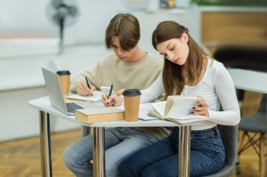 teen girl looking in book and writing near laptop and friend in library