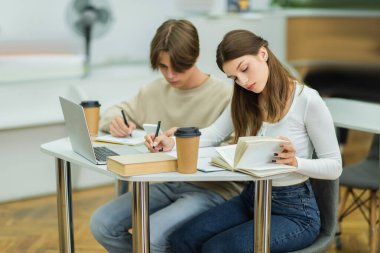 teen girl looking in book and writing near laptop and friend in library clipart
