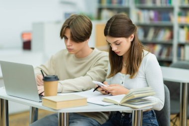 teenage girl using smartphone near student and laptop in library reading room