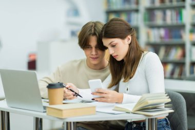 teenage students looking at smartphone near books and laptop in reading room