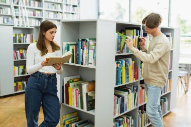 pretty teenage girl reading near friend choosing book in library
