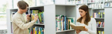 teen guy choosing books on shelves near friend reading in library, banner