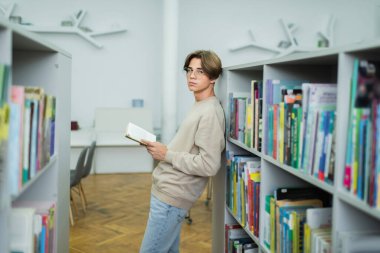 teenage guy in eyeglasses standing with book near racks and looking at camera
