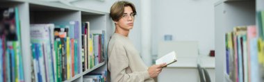 teenage student with book standing near shelves in library and looking at camera, banner