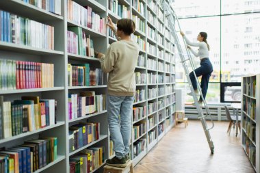 full length of teenage students choosing books on racks in library