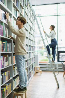 student in eyeglasses choosing books in library near teenage girl on ladder 