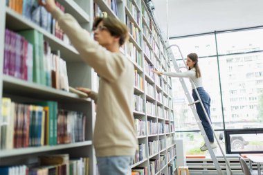 teenage girl standing on ladder and choosing books near friend on blurred foreground