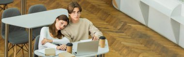 teenage students looking at camera near books and laptop in library, banner