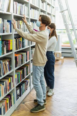 teenagers in medical masks choosing books on racks in library