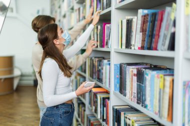 side view of teen girl in medical mask holding smartphone and choosing books near blurred friend
